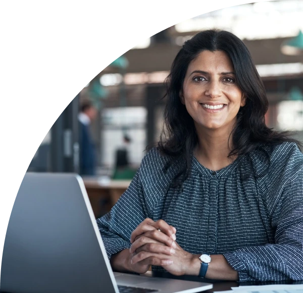 Woman in business casual attire seated at desk behind laptop, smiling into camera