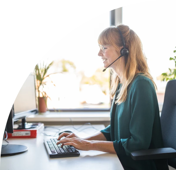Woman wearing business casual attire and telephone headset, seated at desk, working on computer