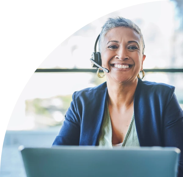 Woman in business casual attire, wearing telephone headset, seated behind laptop and smiling into camera